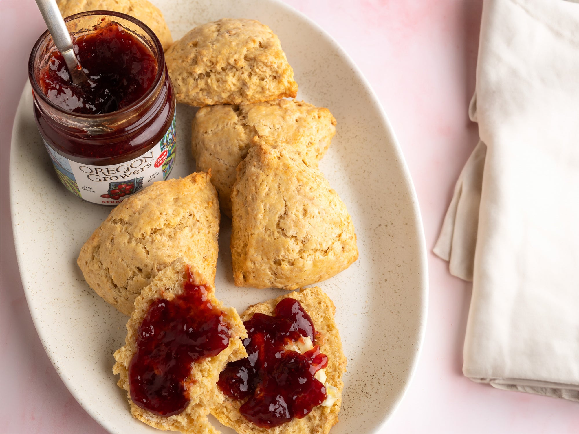 A top view of a plate with Mini Oat Scones covered with Oregon Growers Strawberry Jam and the open jar.