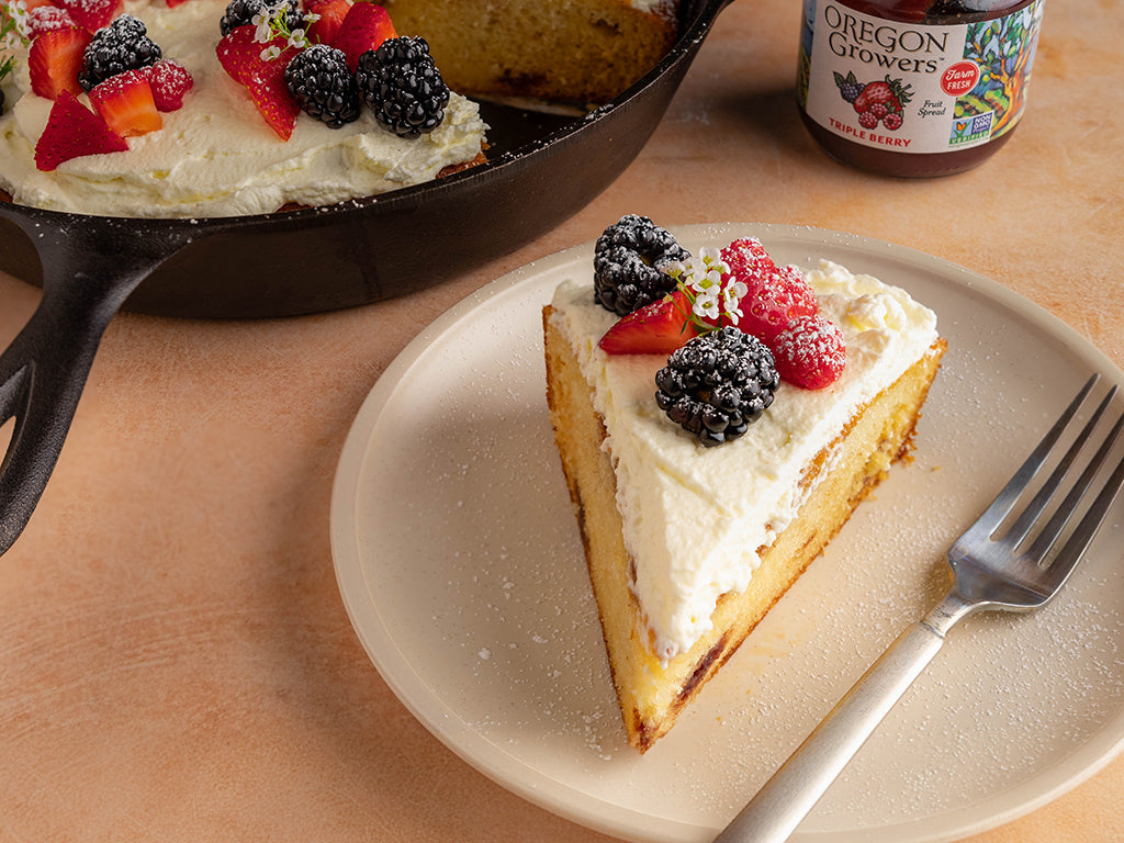 A slice of desert from the Berry Skillet Shortcake recipe, sitting on a plate with a jar Oregon Growers Triple Berry Jam and the serving pan in the background.