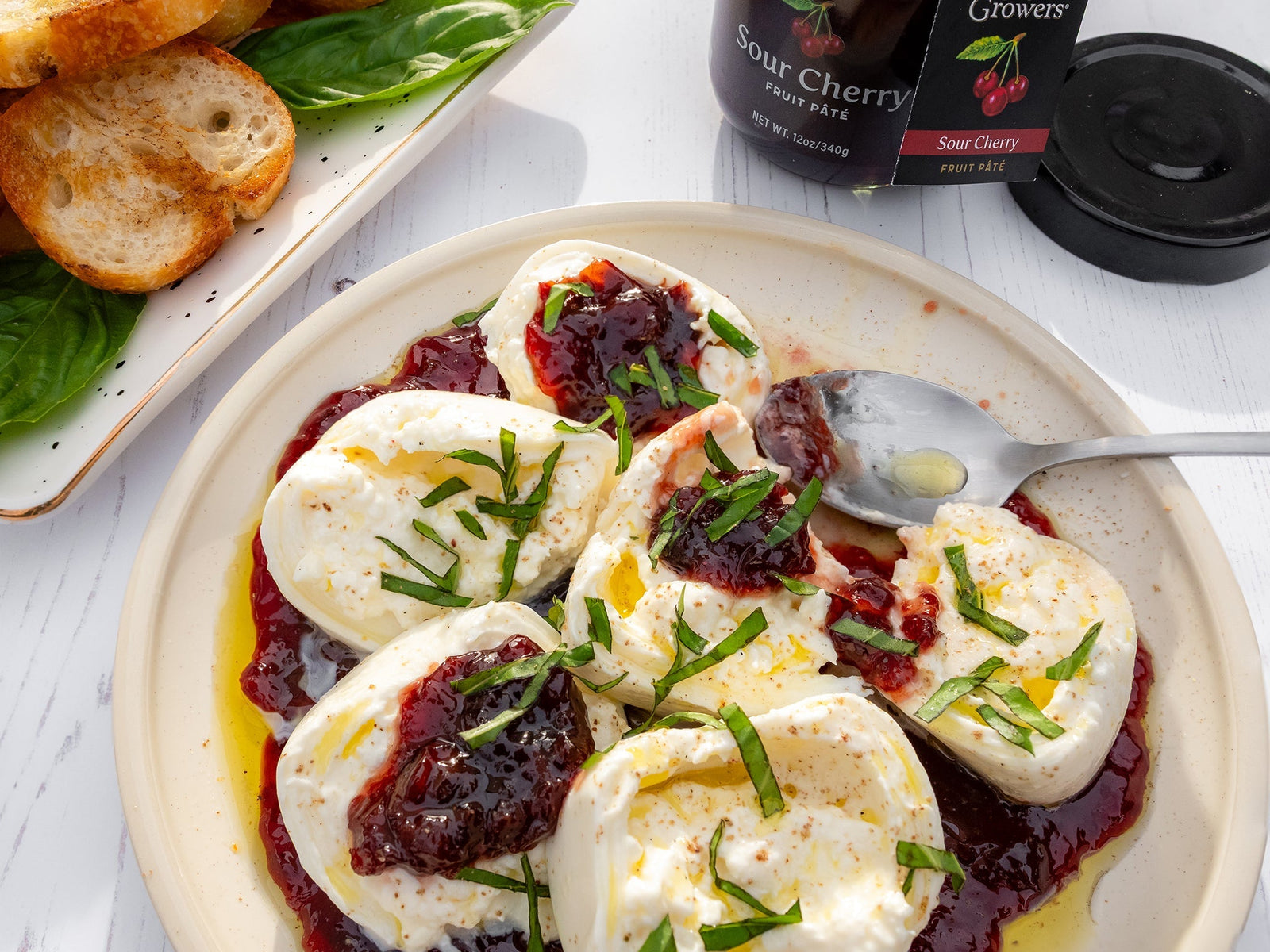 Top view of a plate containing the Burrata With Sour Cherry Fruit Pate next to an opened jar of Oregon Growers Sour Cherry Fruit Pate and a plate of grilled bread