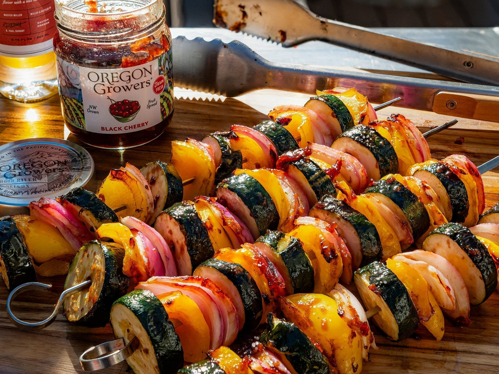 Front view of Cherry Glazed Veggie Kebabs laying on a wooden platter with and opened jar of Oregon Growers Black Cherry Jam and kitchen tongs.