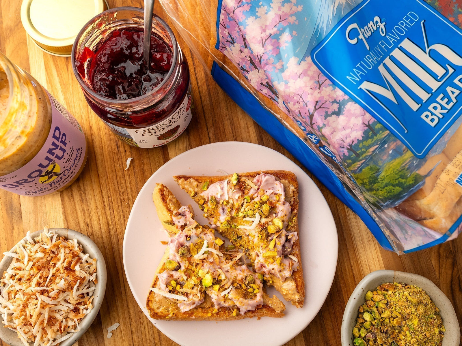 Top view of breakfast setup with the Oregon Growers Cherry Nut Toast recipe, with open jars of Triple Nut Crunch Nut Butter and Black Cherry Jam, and a loaf of Franz Milk Bread on a wooden table.