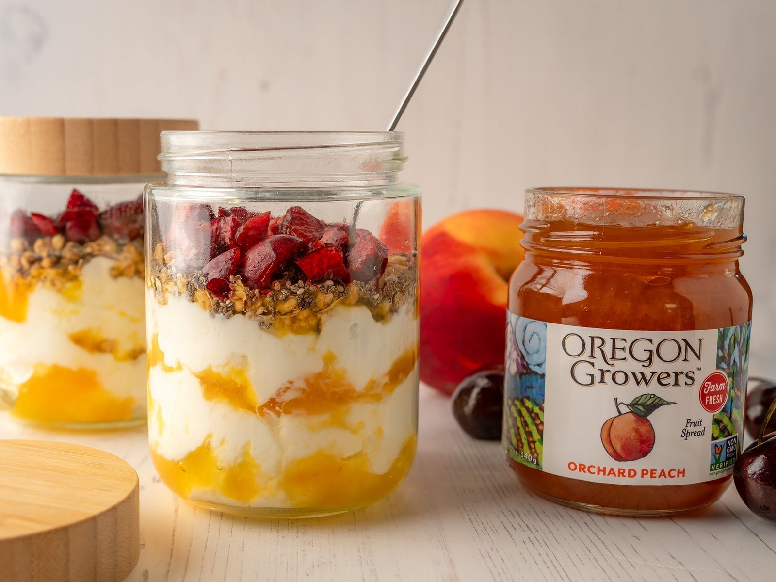 Side view of Cherry Peach Parfait in a jar with spoon, next to opened Oregon Growers Orchard Peach Jam on a table setting, ready to served.
