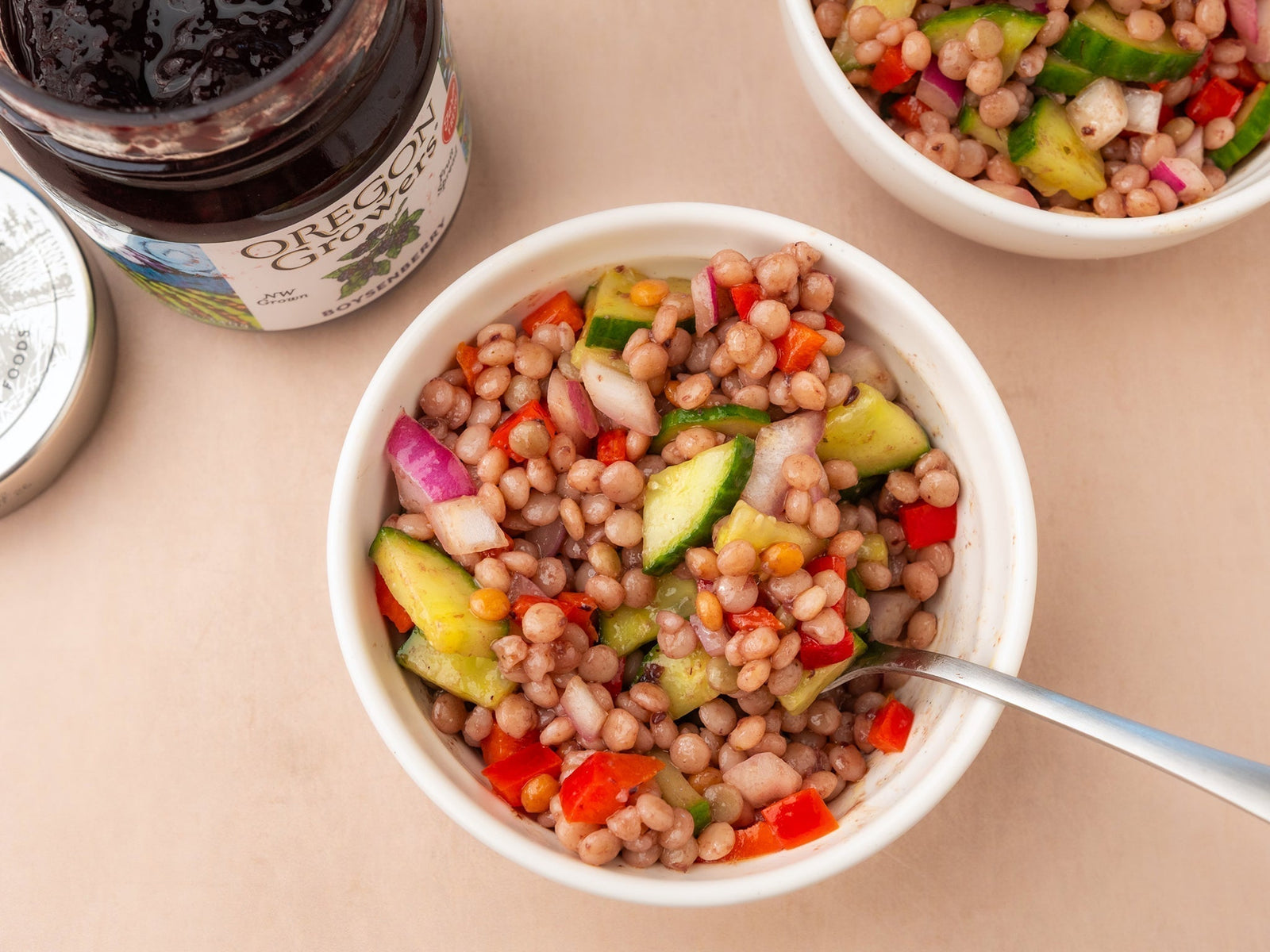Top-down view of Oregon Growers Couscous Salad recipe - pearl couscous with red pepper, red onion, and cucumber - served with an open jar of Oregon Growers Boysenberry Jam beside the bowl.