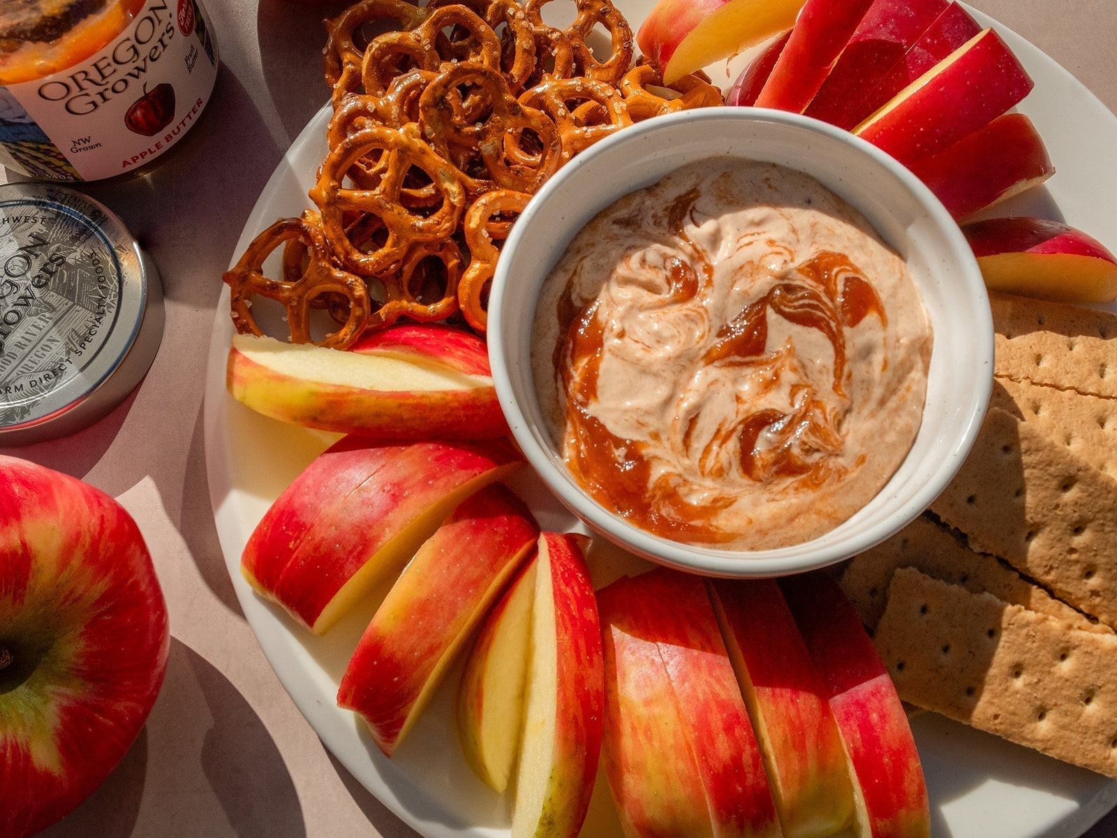 The Easy Apple Butter Dip, a recipe from Oregon Growers, in a bowl surrounded by apples, graham crackers, and pretzels on a plate with an opened jar of Oregon Growers Apply Butter nearby.