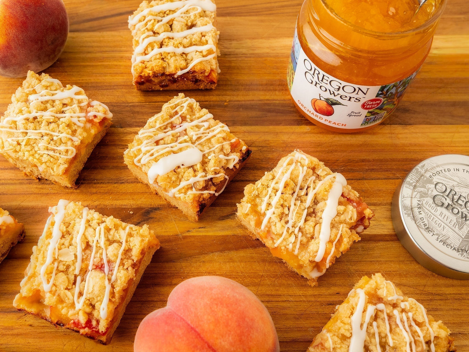Fresh Peach Bars topped with a sweet white drizzle arranged on a wooden surface, with a jar of Oregon Growers peach jam and whole peaches nearby.