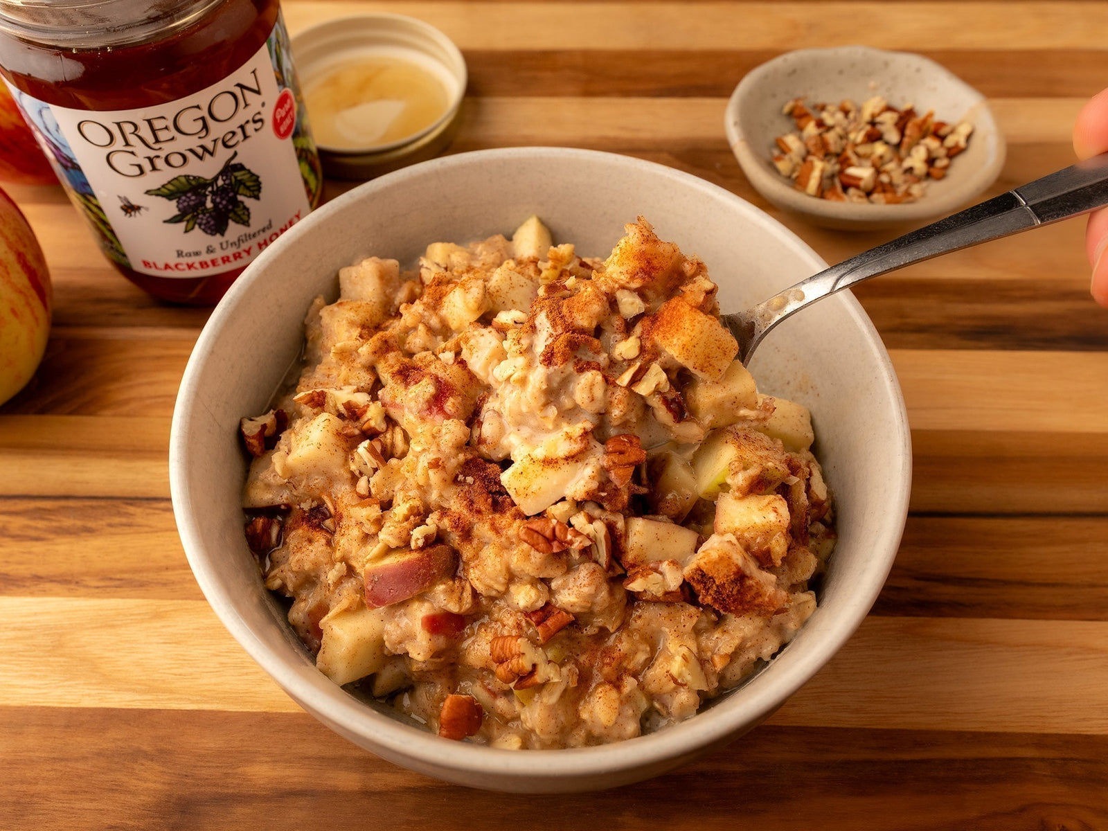A bowl of Oregon Growers Honey-Oat Oatmeal with diced apples, oats, cinnamon, salt, and honey, served with a spoon and a open jar of Oregon Growers Blackberry Honey in the background.