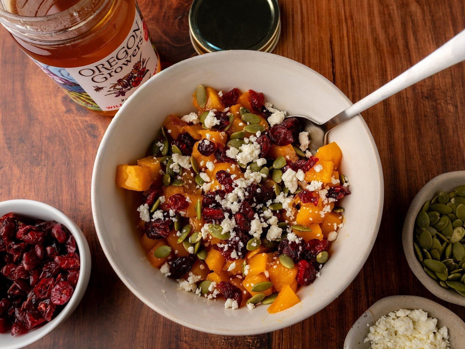 A top view of the Honey Roasted Butternut Squash, a recipe from Oregon Growers, in a bowl surrounded by ramekins of pepitas, dried cranberries, and crumbled cheese, with an opened jar of Oregon Growers Wildflower Honey nearby.