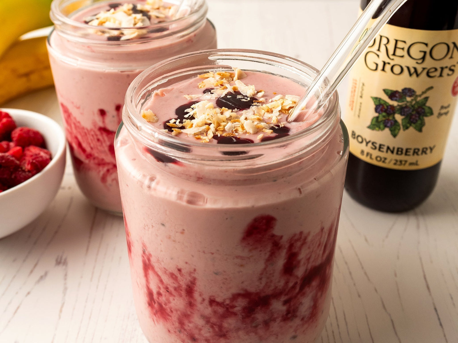 A glass filled the the PB, Banana & Boysenberry Smoothie with fresh bananas, raspberries, and a bottle of Oregon Growers Boysenberry Fruit Syrup in the background.