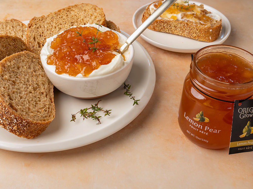 A side view of a serving plate containing bread slices and a bowl of Pear & Blue Cheese Dip. Next to a jar of Oregon Growers Lemon Pear Fruit Pate.