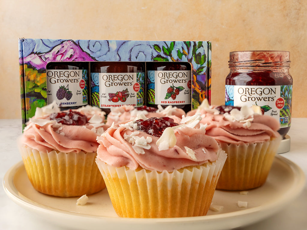 A plate of three Raspberry Coconut Cupcakes in front of a jar of Oregon Growers Red Raspberry Jam and the Oregon Berry Gift Trio gift box.
