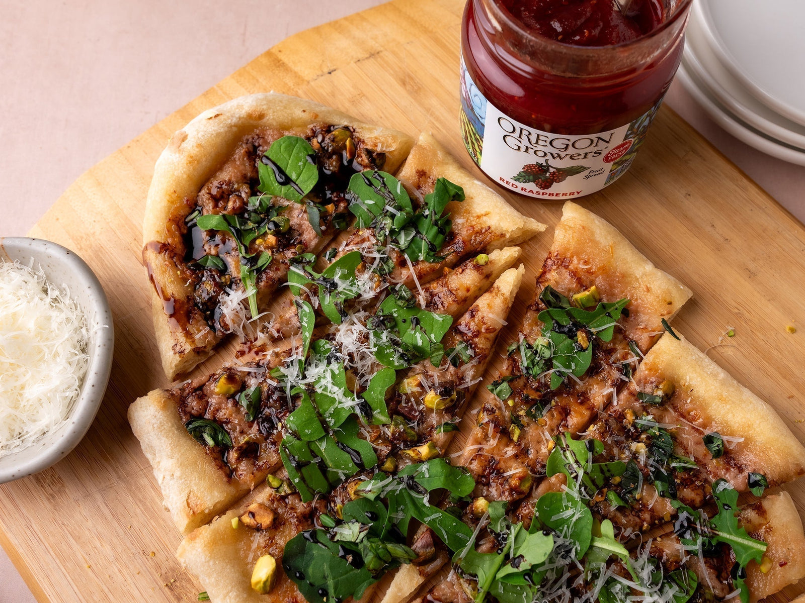 Top View of the Raspberry, Pistachio & Arugula Flatbread with toppings on a wooden serving platter next to a jar of Oregon Growers Red Raspberry Jam and a bowl of cheese.