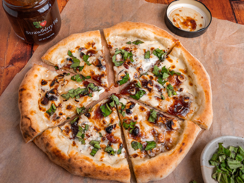 Top view of the Oregon Growers Strawberry Fig Dessert Pizza cut in slices with an open jar of Starwberry Fig Pate and a bowl of minced Basil on the side.