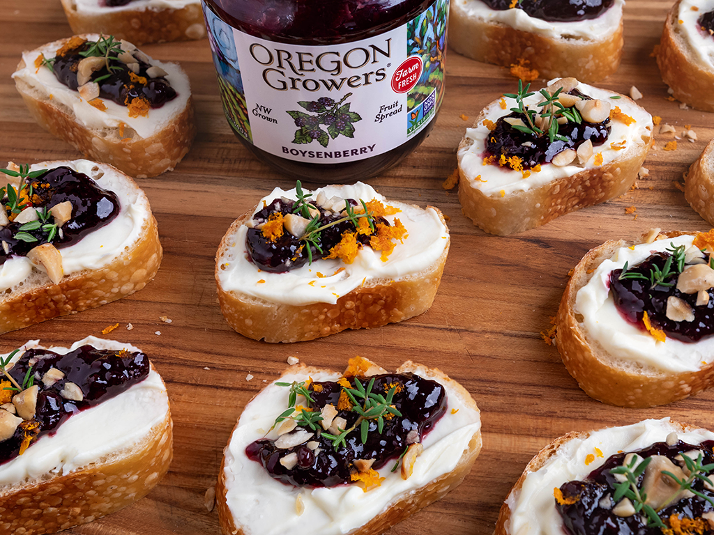 A tabletop view with a jar of Oregon Growers Boysenberry Jam in front of slices of Boysenberry Crostini Bites scattered evenly on the table.