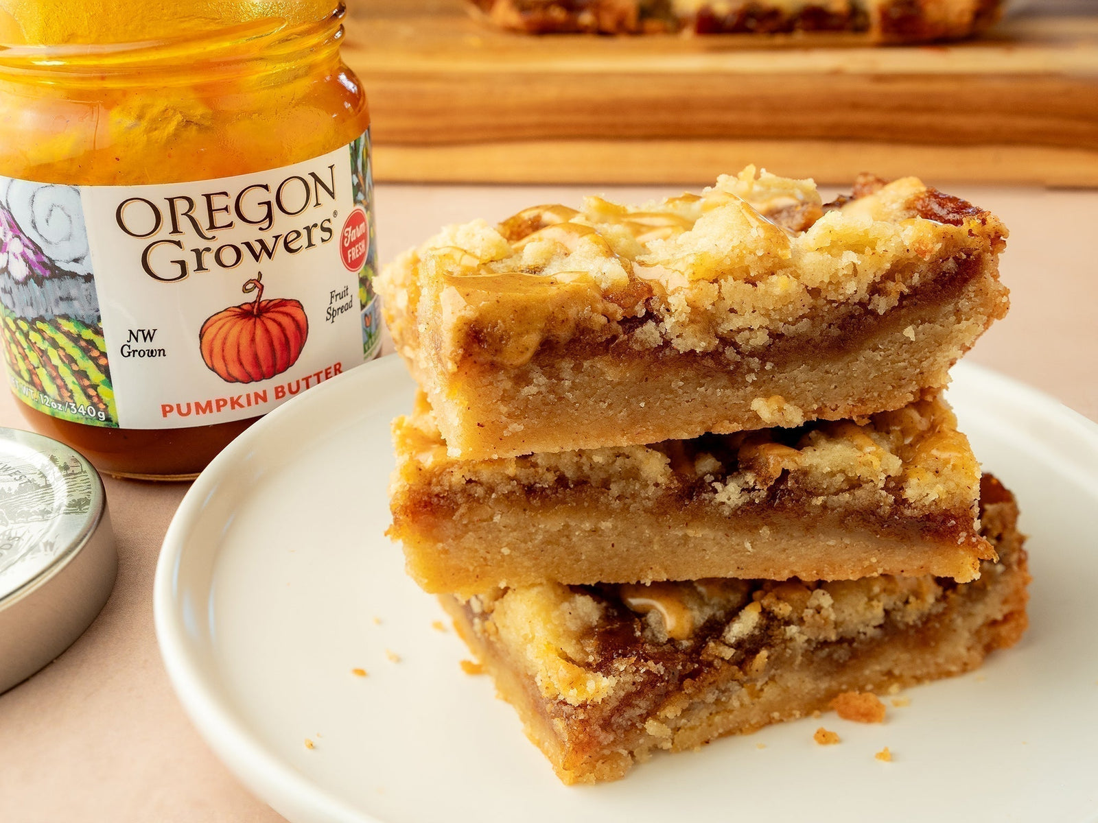Three stacked Gooey Pumpkin Butter Bars on a white plate, with a jar of Oregon Growers pumpkin butter and a wooden cutting board in the background.
