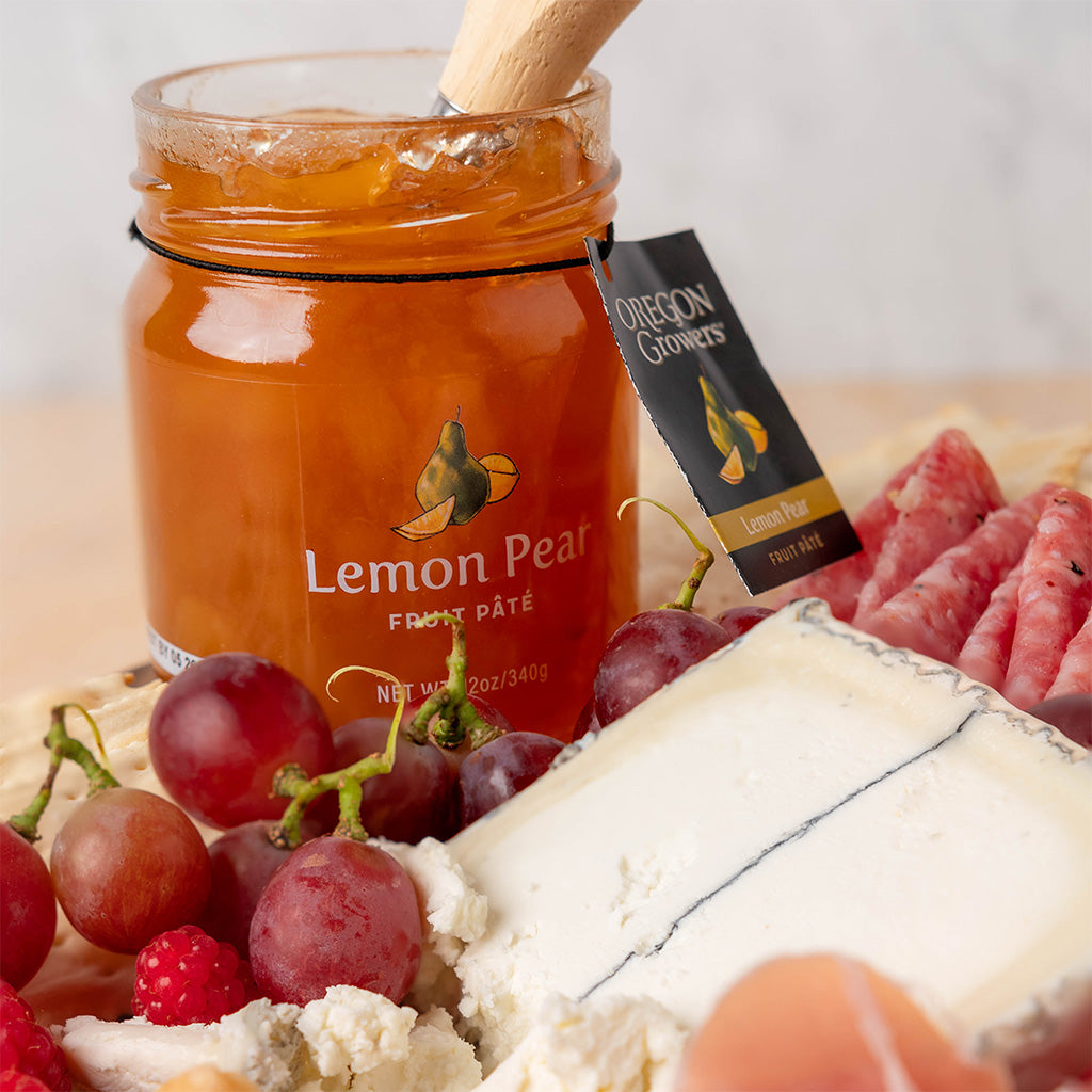 Close-up of Oregon Growers Lemon Pear Pate jar in front of fruit, cheese, and other snacks.